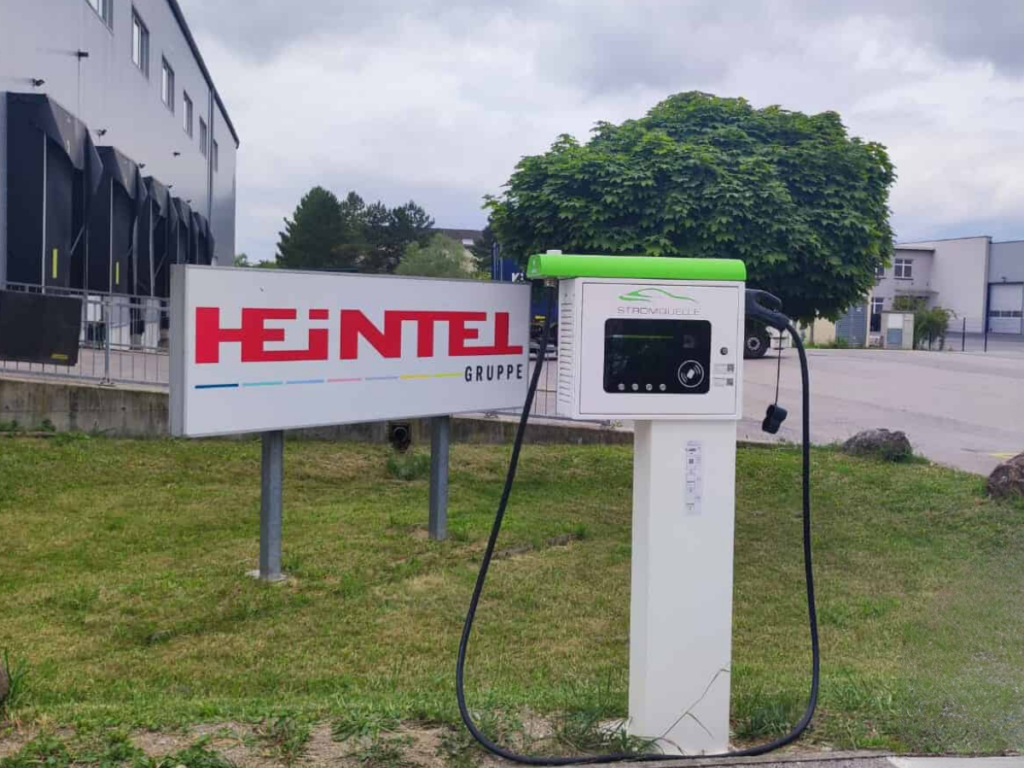 Stromquelle-Ladestation mit grünem Dach vor einem Firmenschild der HEINTEL Gruppe. Die Ladesäule steht auf einer Grünfläche neben einem Industriegebäude und ist mit einem Display und einem Ladekabel ausgestattet. # Ejoin Rapid Box