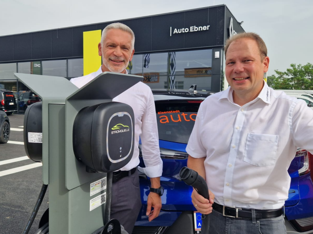 Zwei Männer in weißen Hemden stehen neben einer Stromquelle-Ladestation auf dem Gelände von Auto Ebner. Einer hält ein Ladekabel in der Hand, während im Hintergrund ein blaues Elektrofahrzeug und das Firmengebäude zu sehen sind. #Wallbox Pulsar Pro