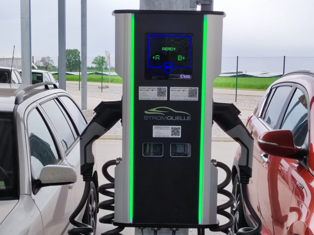 Eine Stromquelle-Schnellladestation mit zwei angeschlossenen Ladekabeln lädt gleichzeitig zwei Elektrofahrzeuge unter einer überdachten Parkfläche auf. Die Ladestation ist mit einem digitalen Display ausgestattet und zeigt den Ladestatus an. Elita CityCharge Mini