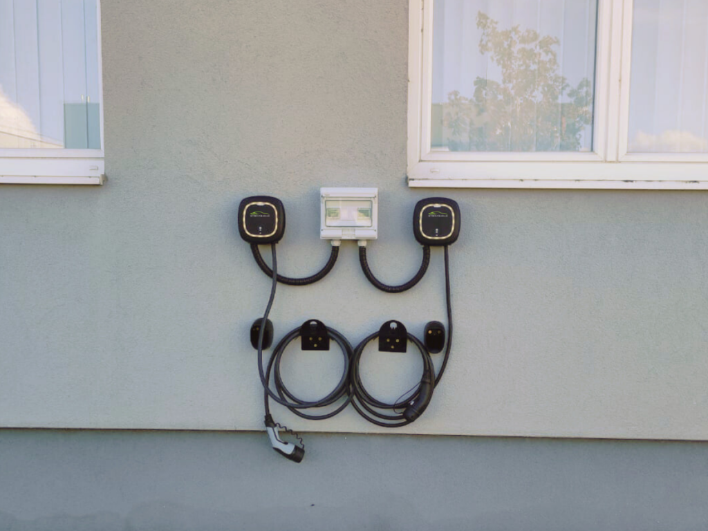 Zwei kompakte Stromquelle-Wandladestationen an einer Hauswand unter Fenstern, ausgestattet mit Ladekabeln und einer zentralen Steuereinheit. Die Ladekabel sind ordentlich auf Halterungen aufgerollt. #Wallbox Pulsar Pro