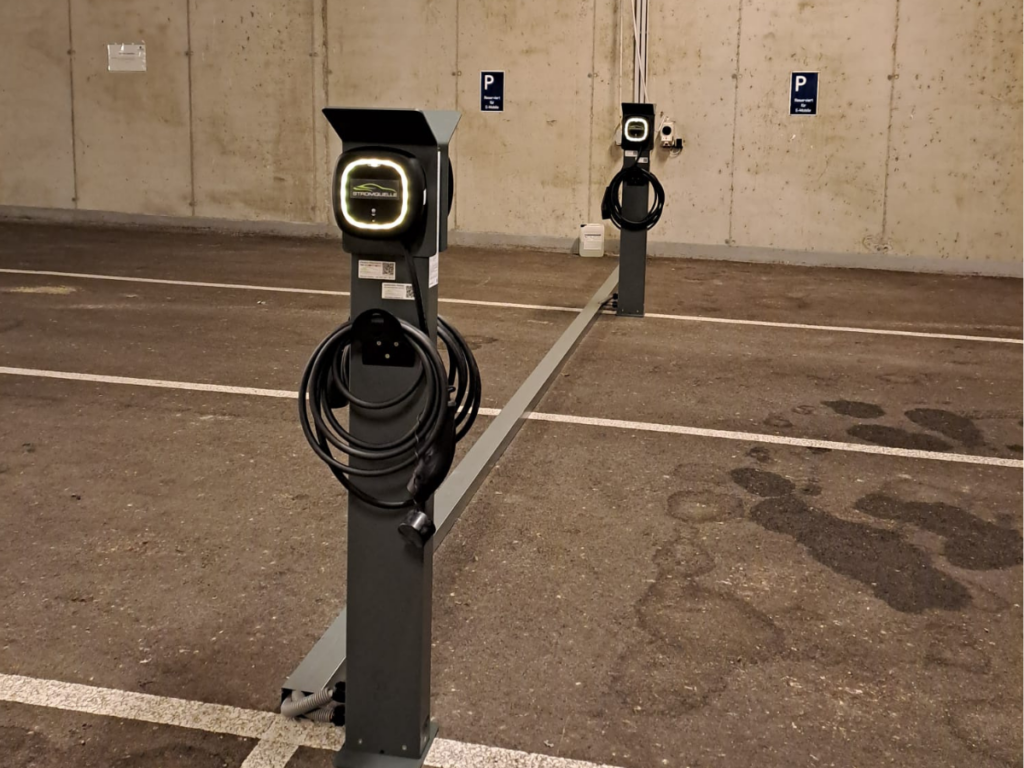 Zwei Stromquelle-Ladestationen in einer Tiefgarage mit markierten Parkplätzen für Elektrofahrzeuge. Die Ladestationen sind mit einem beleuchteten Logo und einem aufgerollten Ladekabel ausgestattet. #Wallbox # Pulsar Pro